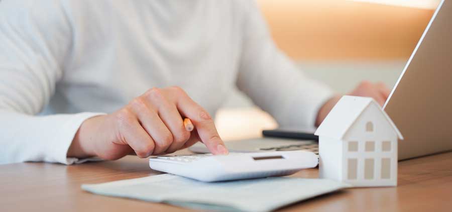 A person with a pencil, notepad, and calculator in front of a computer and miniature house model
