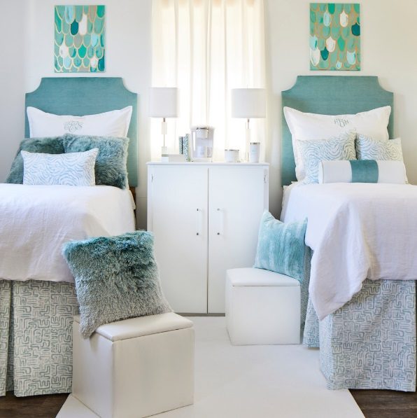 Front facing view of two decorated twin size beds on alternate sides, with a white cabinet in between as a night stand and a small lamp on each side