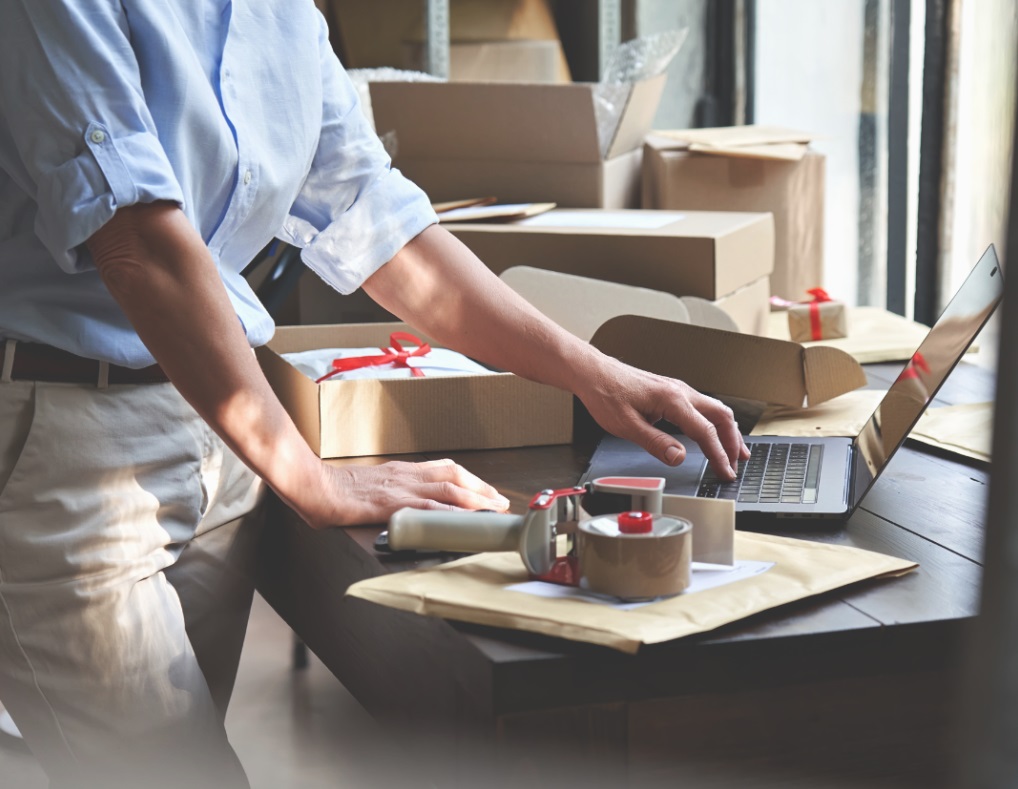Person standing at a table, looking at a laptop and surrounded by packing supplies