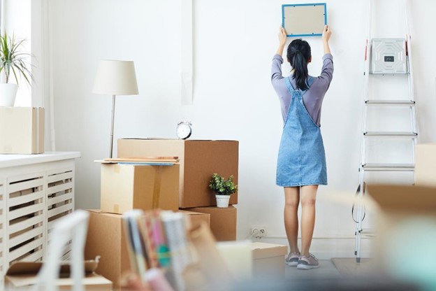 a college student is surrounded by moving boxes while she tries to hang a picture on the wall