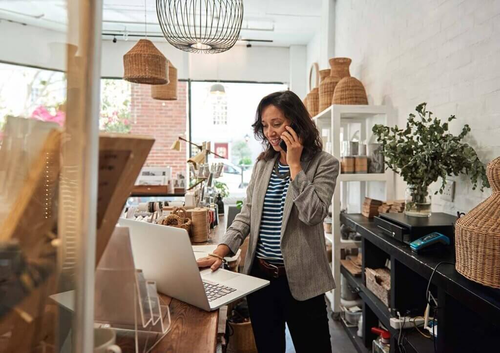 Female shop owner standing behind counter while speaking on the phone and looking at her laptop computer