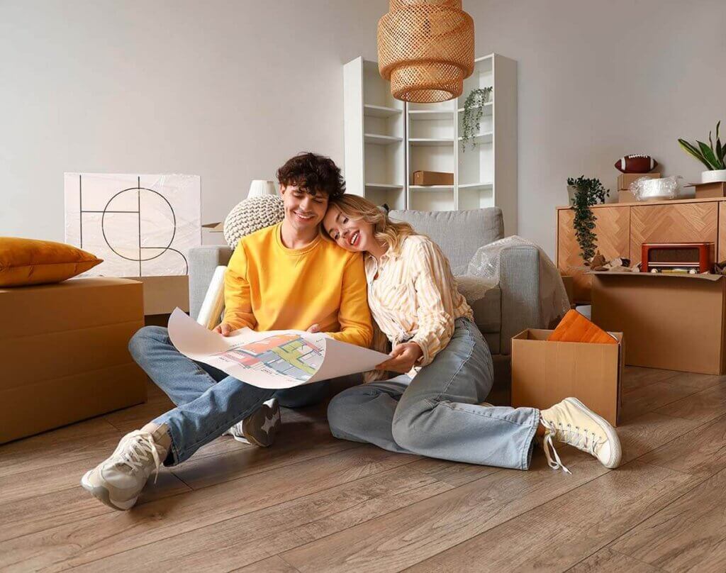 Couple sitting on the floor of their new home, each holding one end of a blueprint.